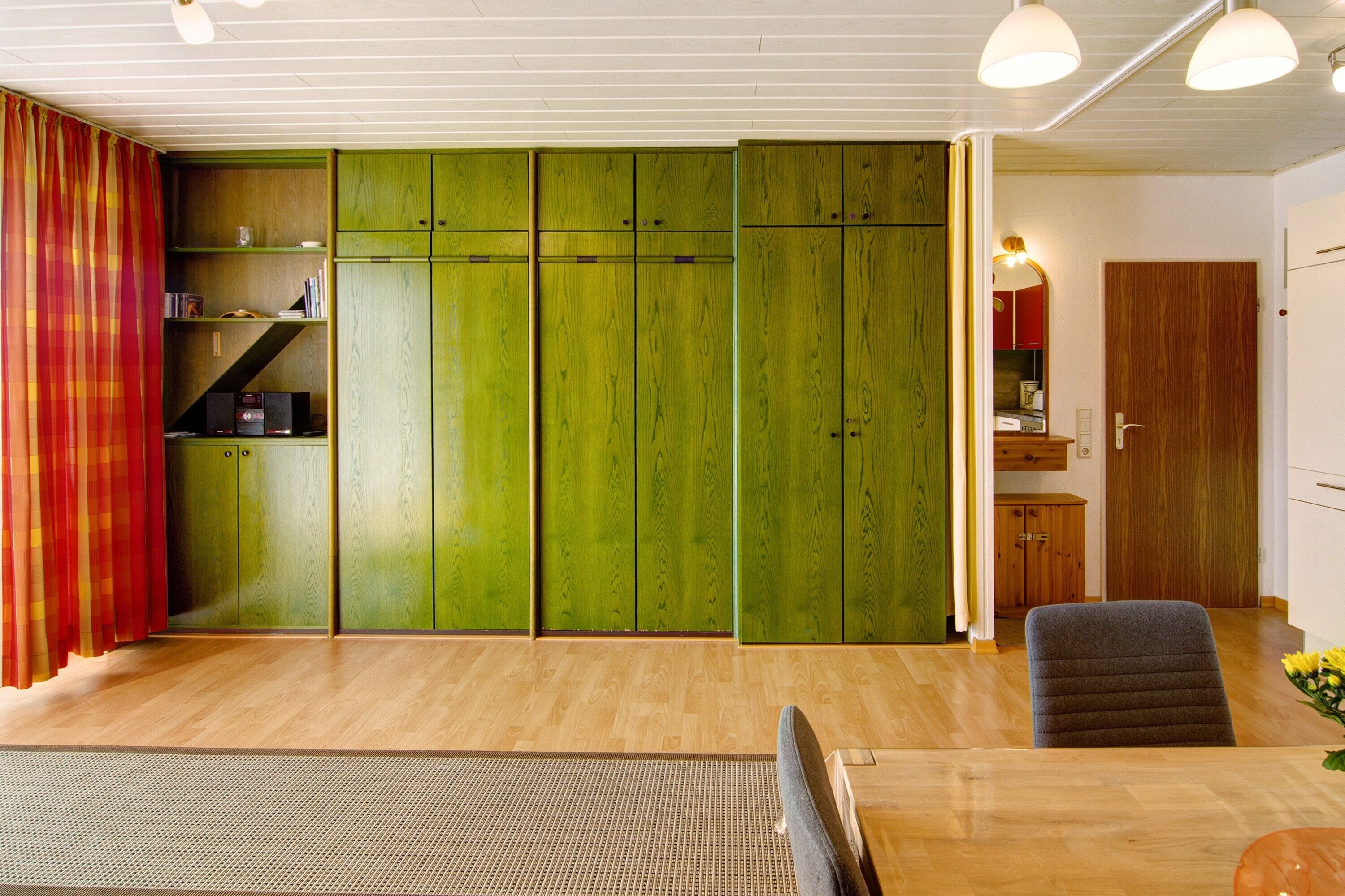 Una habitación con piso de madera, una mesa de comedor con sillas, un armario verde y una zona de cocina con armarios.