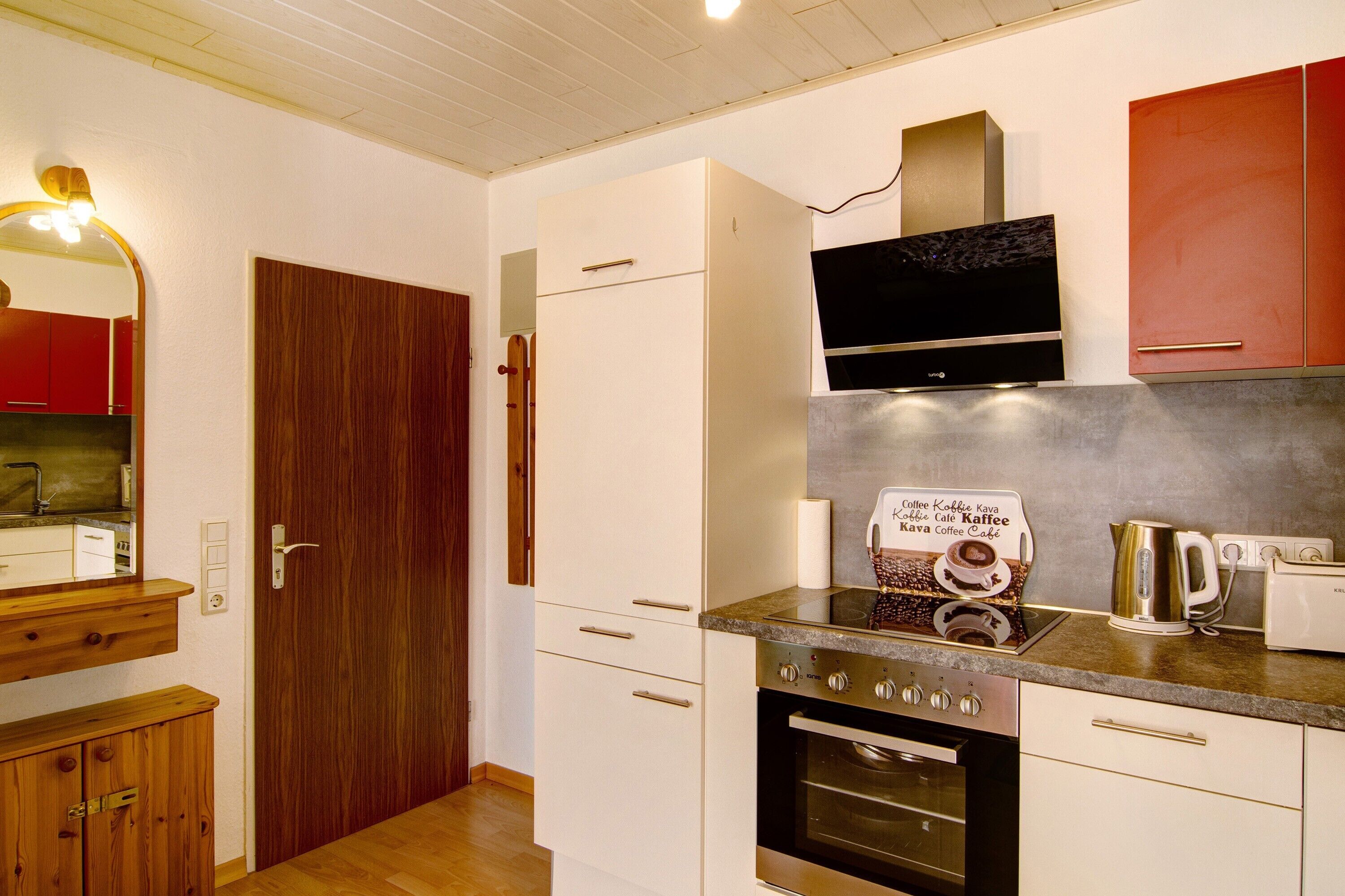 A modern kitchen with white cabinets, a wooden door, a built-in oven, and a coffee maker.