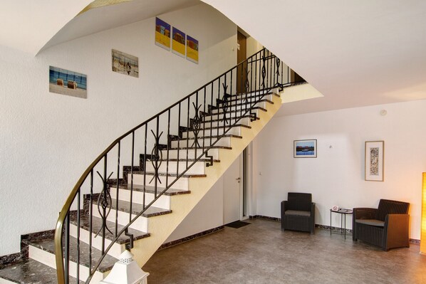Una escalera con barandilla negra, una puerta blanca y dos sillas de mimbre en una habitación con suelo de baldosas.