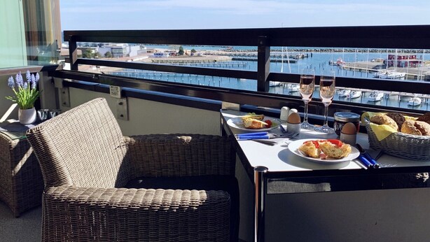 Un balcón con mobiliario de mimbre, una mesa con comida y bebidas, y vista a un puerto deportivo.
