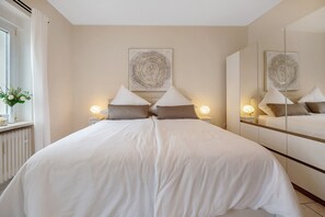 Un lit double avec une couette blanche, deux lampes de chevet, un grand miroir et une œuvre d’art accrochée au mur.