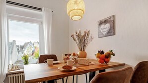 Un comedor con una mesa de madera, sillas, un jarrón con flores secas, un frutero y una señal colgada en la pared.