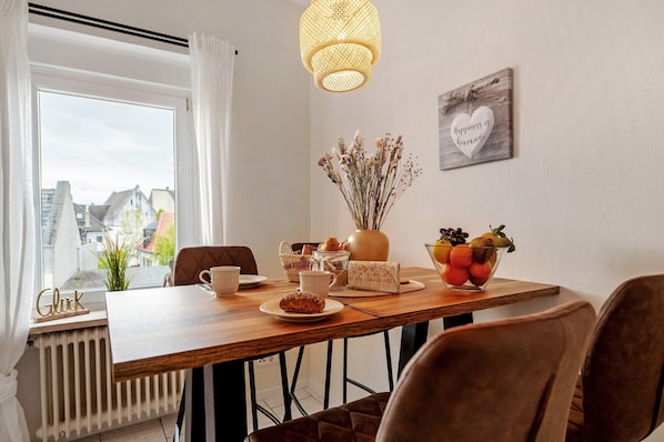 Un coin repas avec une table en bois, des chaises, un vase contenant des fleurs séchées, un saladier de fruits et un panneau fixé au mur.