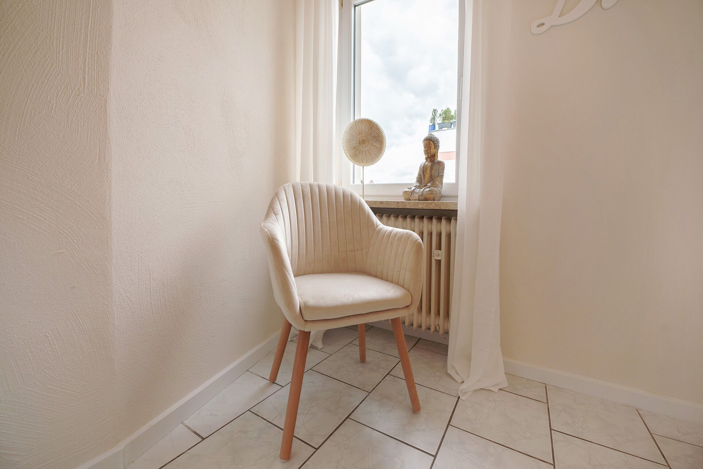 Ein Zimmer mit Stuhl, Fenster mit Ausblick, Heizkörper und Wandventilator.
