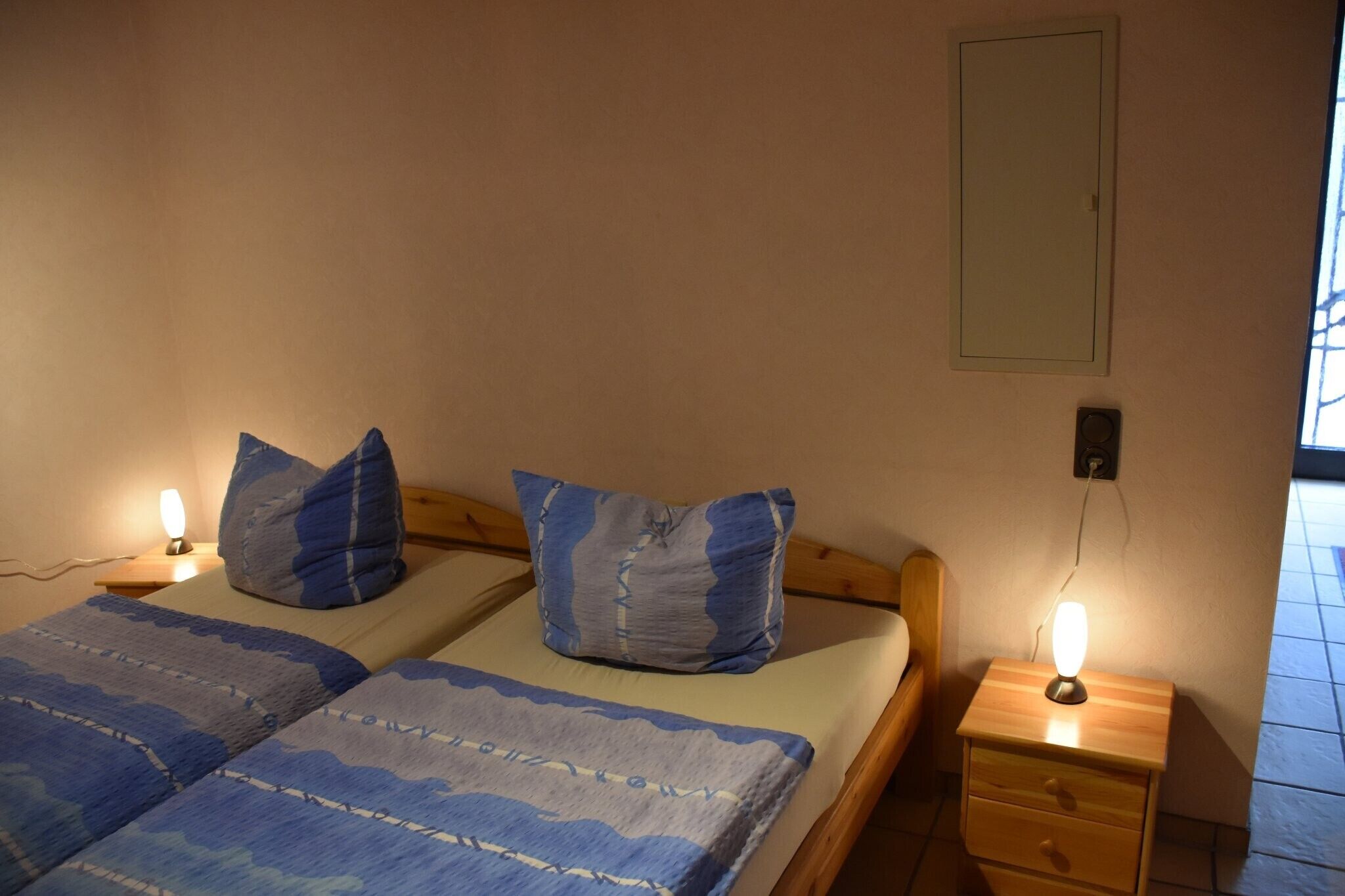Un lit double avec une literie bleue, deux oreillers, des tables de chevet avec des lampes et un interrupteur fixé au mur.