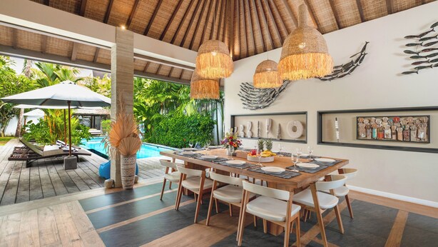 A dining area with a wooden table set for six, surrounded by white chairs, under a thatched ceiling. - Villa Nikaia by Villa Finder (Seminyak)