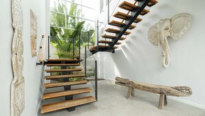 A modern staircase with wooden steps and black metal railings, leading to a room with a large window and tropical plants. - Villa Nikaia by Villa Finder (Seminyak)