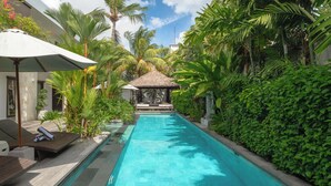 A swimming pool surrounded by lush greenery, a thatched-roof gazebo, and lounge chairs. - Villa Nikaia by Villa Finder (Seminyak)