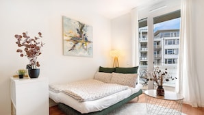 A bedroom with a bed, a side table, a lamp, a window with a view of buildings, and a painting on the wall.