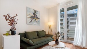 A modern living room with a green sofa, a white coffee table, a painting on the wall, and a view through a glass door.