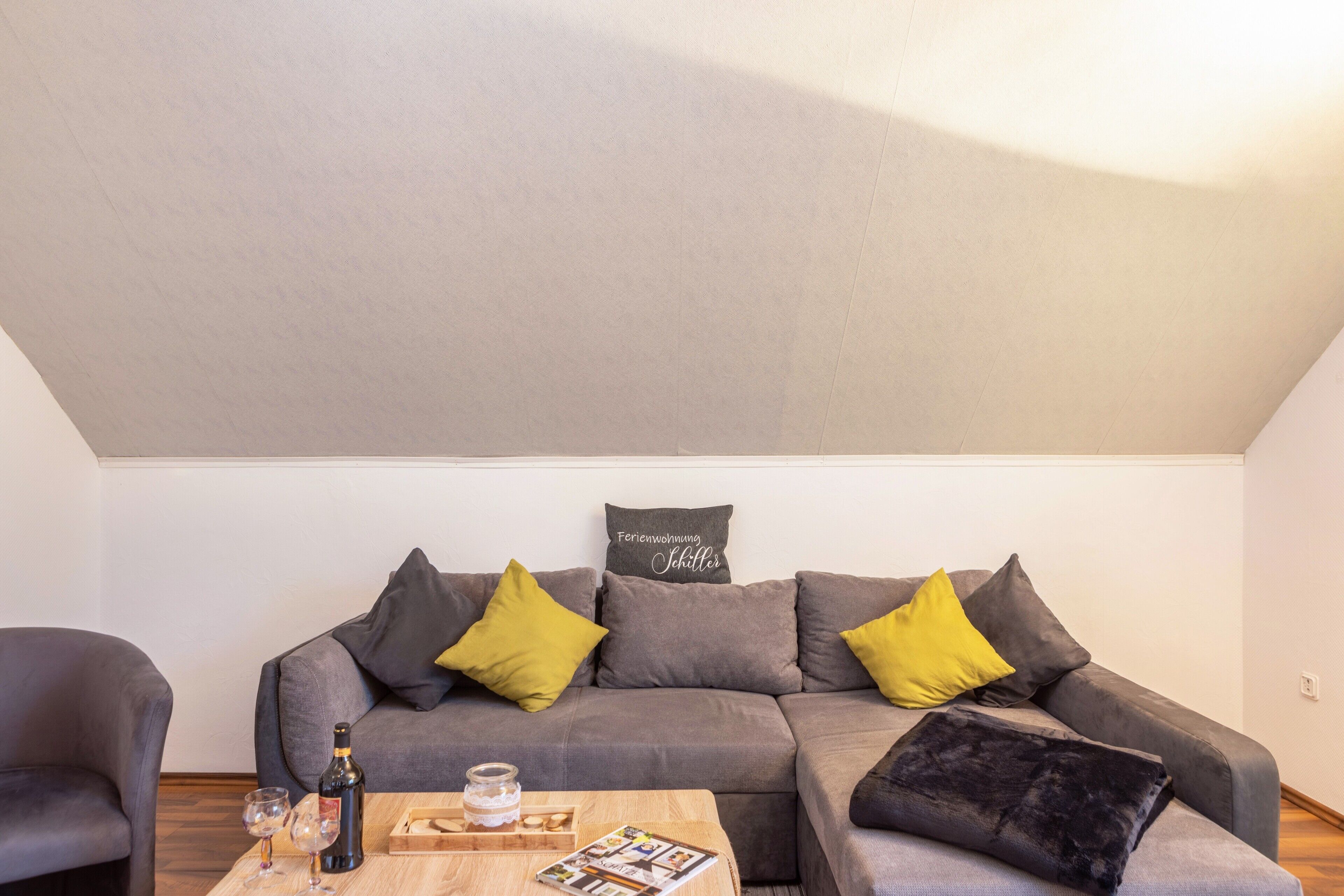 Ein modernes Wohnzimmer mit einem grauen Sofa, gelb-grauen Kissen, einem Holztisch mit Flasche und Gläsern und einem gerahmten Schild an der Wand.