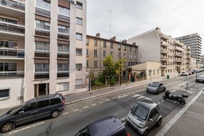 Un immeuble résidentiel à plusieurs étages doté de balcons, une rue bordée de voitures garées et un ciel dégagé.