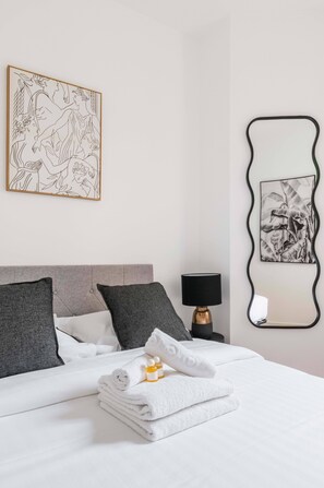 A neatly made bed with white linens, a folded towel, and a bottle of lotion. There is a black lamp on a bedside table, a framed abstract artwork on the wall, and a mirror with a unique frame. - Modern 60m² Apartment Near Metro - Paris 12 (Paris)