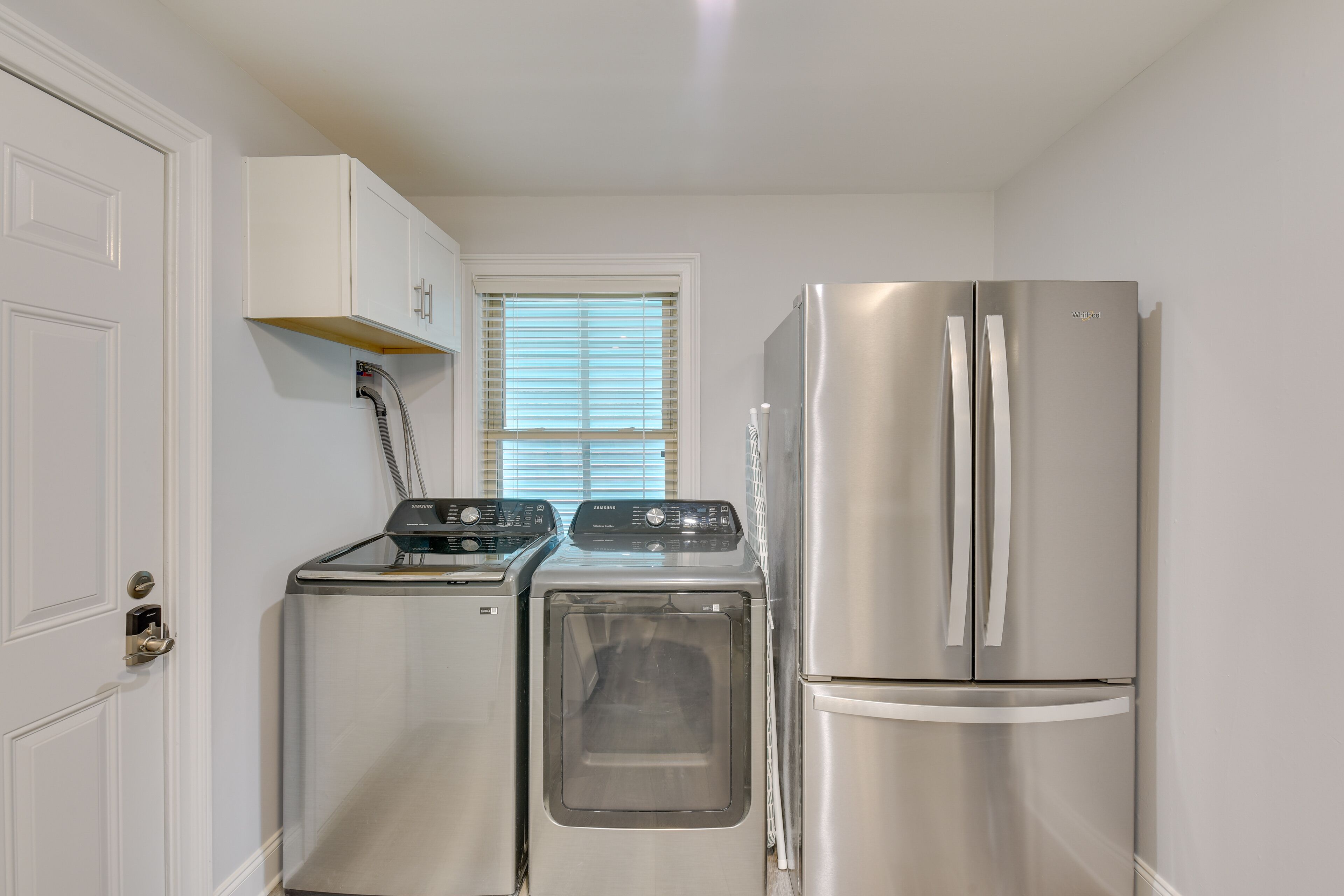 A compact kitchen with a stainless steel washer and dryer, a refrigerator, and a white cabinet above the dryer.