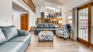 Ein geräumiger Wohnbereich mit einem grauen Sofa, einem gemusterten Hocker und einem Karosessel. Eine Holztreppe führt auf eine höhere Ebene.