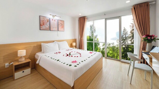 Ein Hotelzimmer mit Bett, Nachttisch, Lampe, Schreibtisch, Stuhl und einem großen Fenster mit Vorhängen.