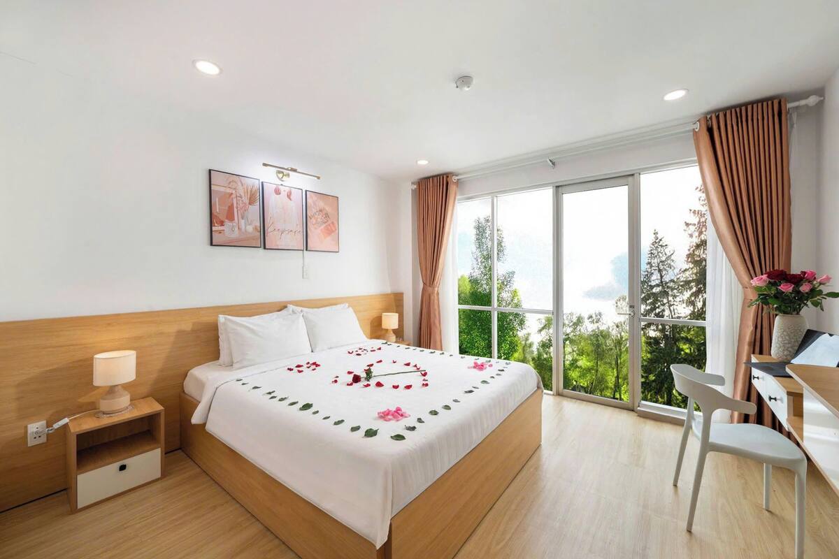 Habitación de hotel con cama, mesita de noche, lámpara, escritorio, silla y ventana grande con cortinas.