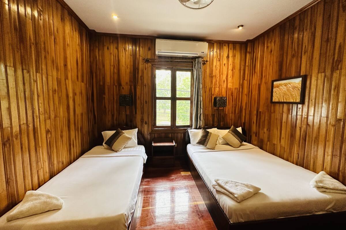 A room with wooden paneling, two beds with white linens, a ceiling fan, and a window with a view of greenery.