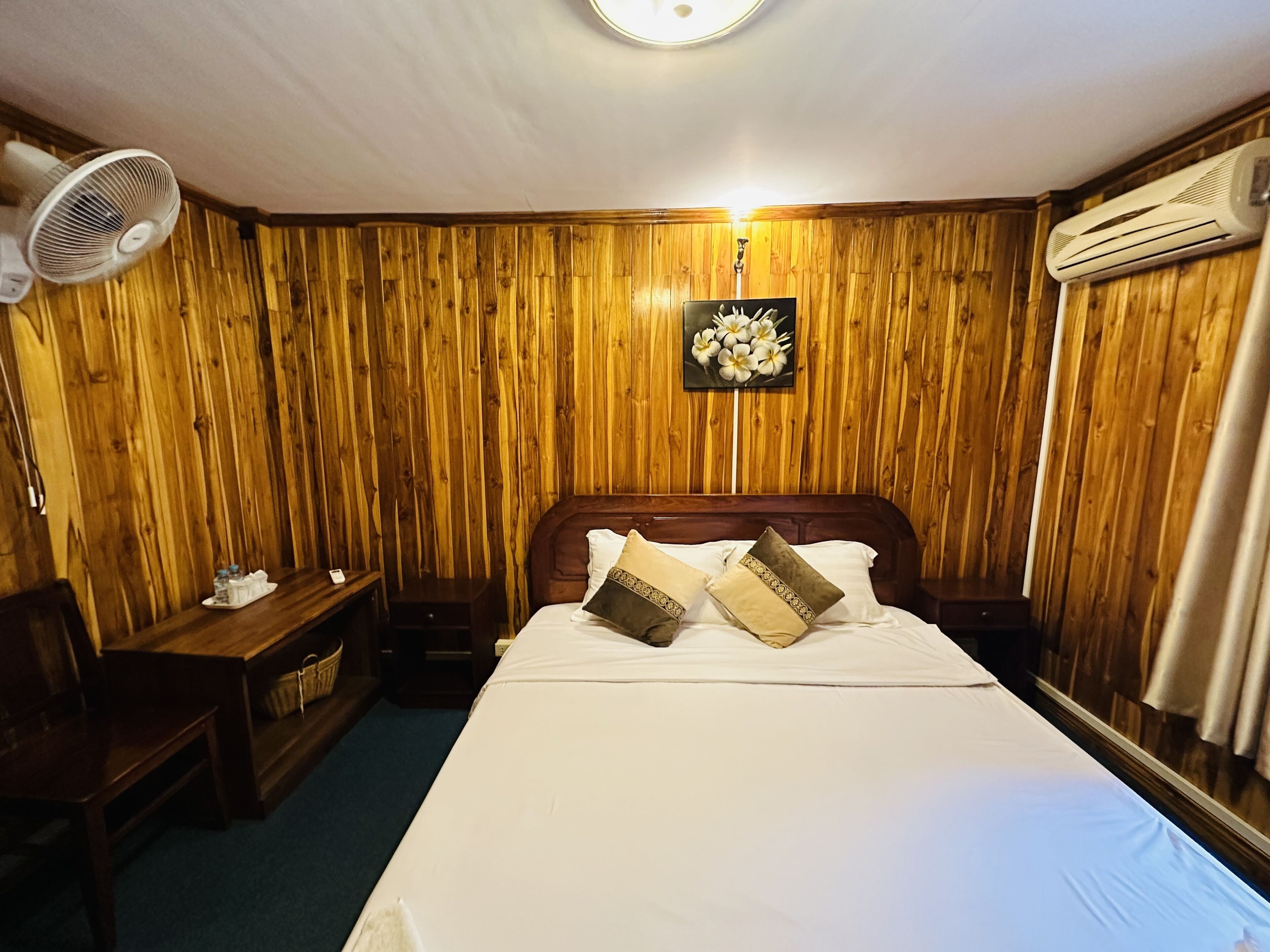 Ein Zimmer mit einer holzverkleideten Wand, einem Bett mit weißer Bettwäsche und dekorativen Kissen, einem Nachttisch, einem Stuhl, einem Standventilator und einer Klimaanlage.
