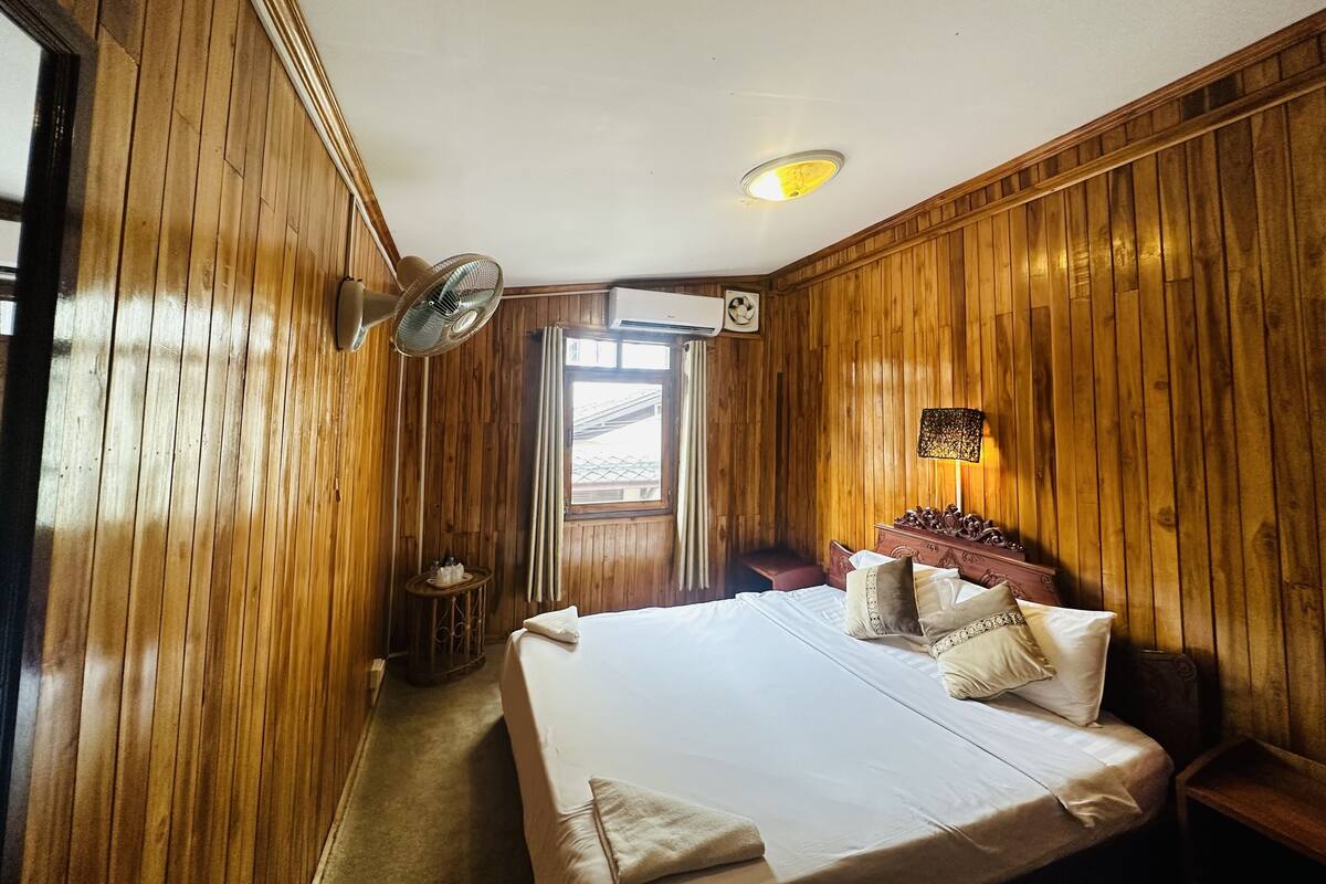 A hotel room with wooden paneling, a bed with white linens, a bedside lamp, and an air conditioning unit.