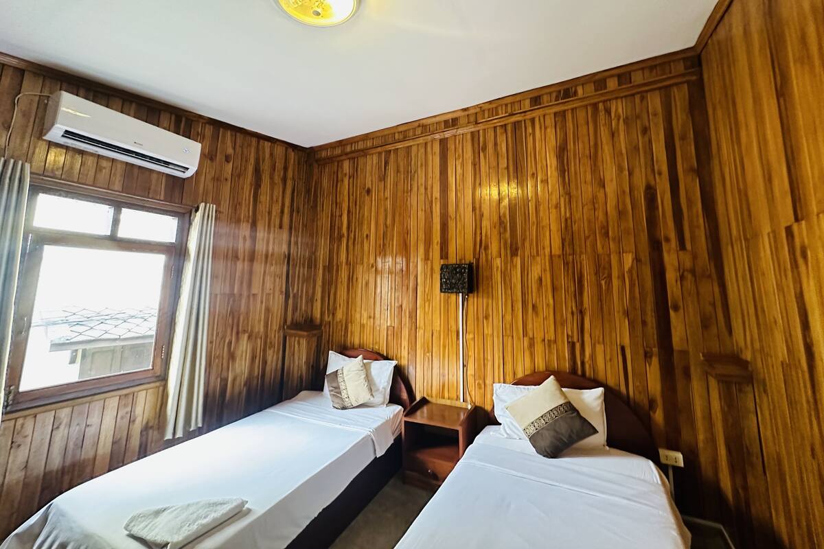 A room with wooden paneling, two beds with white linens, an air conditioning unit, a window with curtains, and a wall-mounted light fixture.