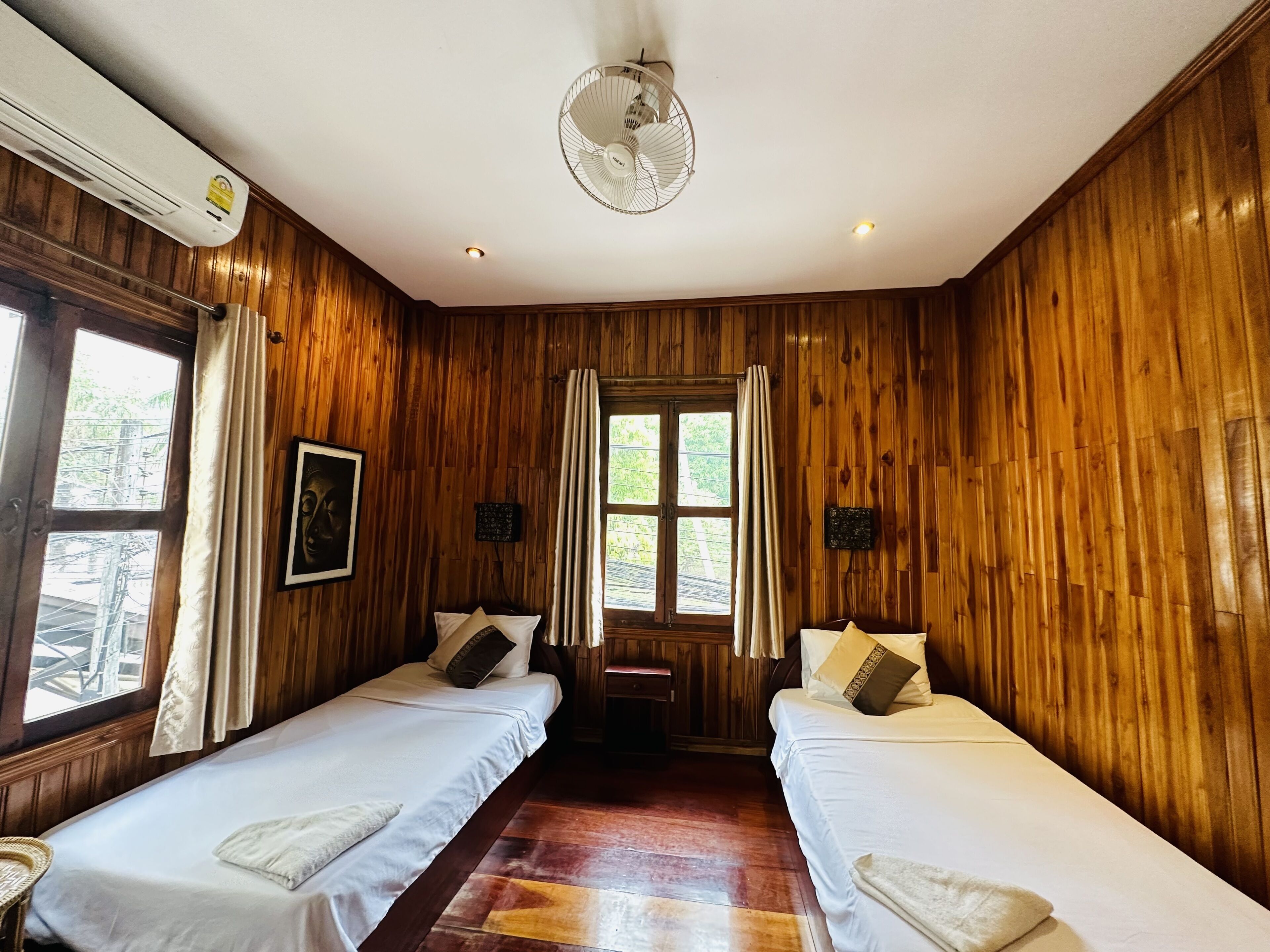 Quarto com paredes de madeira, duas camas com roupa de cama branca, um ventilador de teto e uma janela com cortinas.