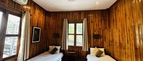 Quarto com paredes de madeira, duas camas com roupa de cama branca, um ventilador de teto e uma janela com cortinas.