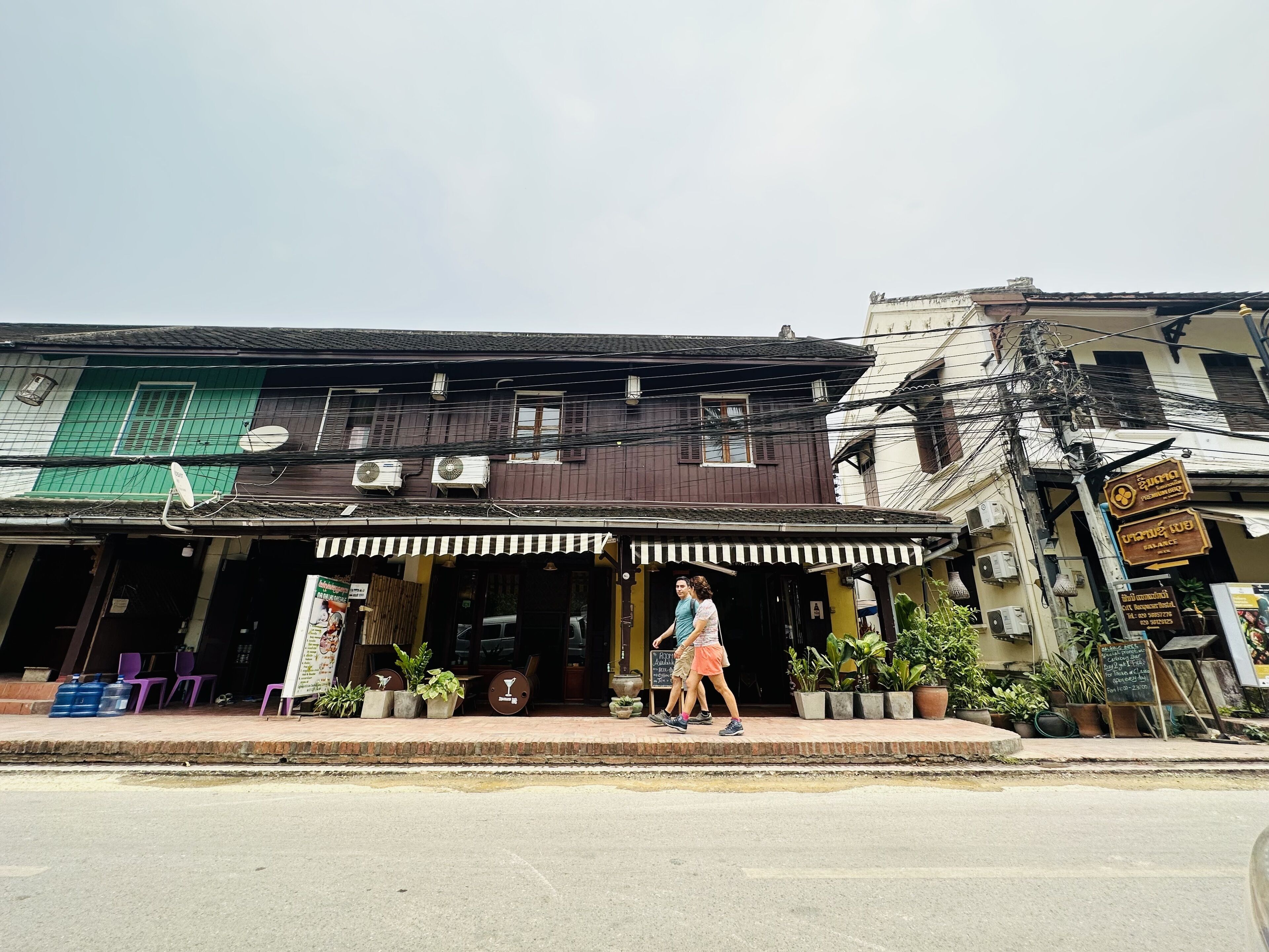 A street view with a building featuring a shop on the ground floor and a restaurant with outdoor seating. There are potted plants and a signboard visible.
