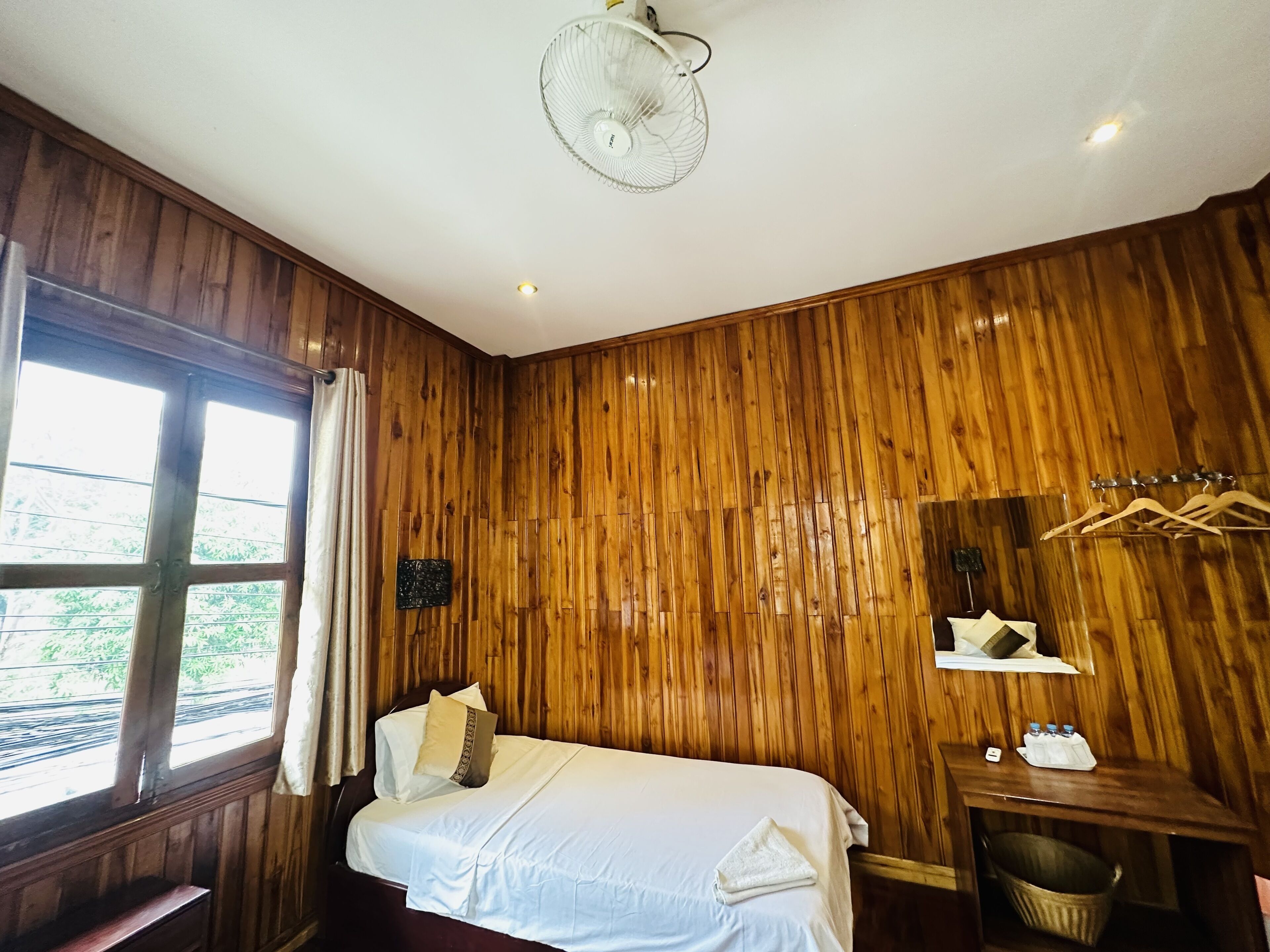 Una habitación con paneles de madera, una cama, un ventilador, una ventana con cortinas, una mesita con una cesta y un espejo con perchero para toallas.