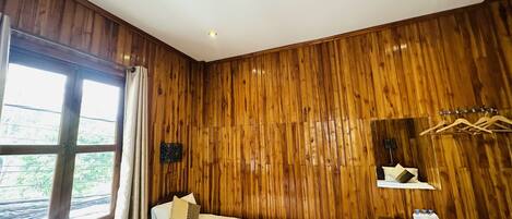 Una habitación con paneles de madera, una cama, un ventilador, una ventana con cortinas, una mesita con una cesta y un espejo con perchero para toallas.