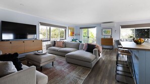 A spacious living room with a large flat-screen TV, a sectional sofa, a coffee table, and a kitchen area with a bar stool and a fruit bowl.