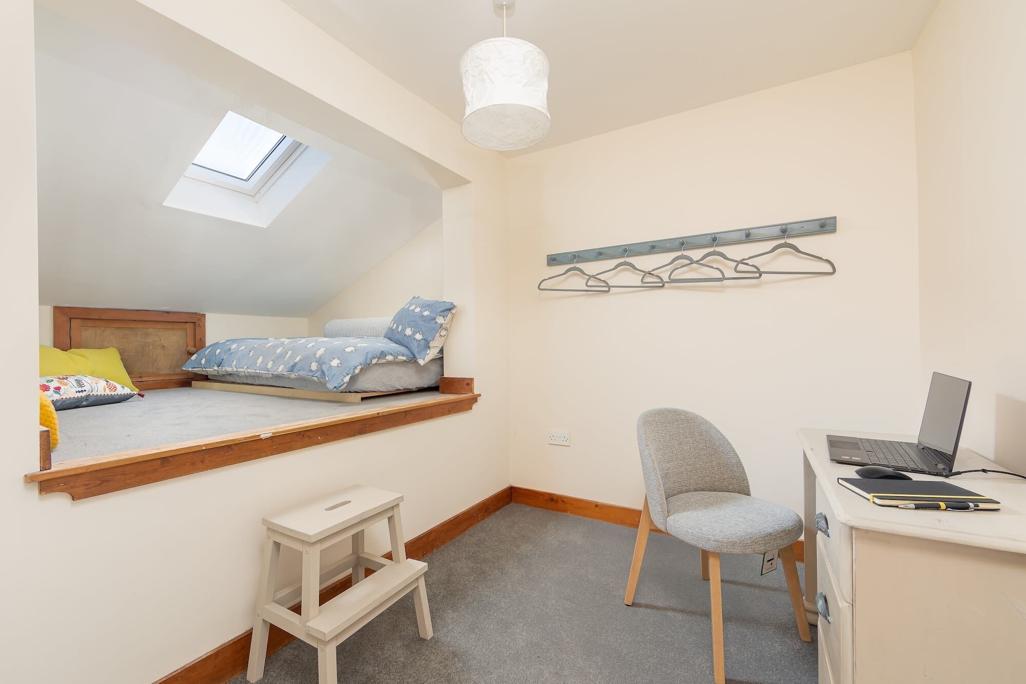 A bedroom with a loft bed, a desk with a laptop, a chair, a small stool, and a wall-mounted coat rack.
