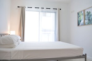 Un lit avec une literie blanche, une fenêtre avec des rideaux légers, une lampe de chevet et une plante décorative accrochée au mur.