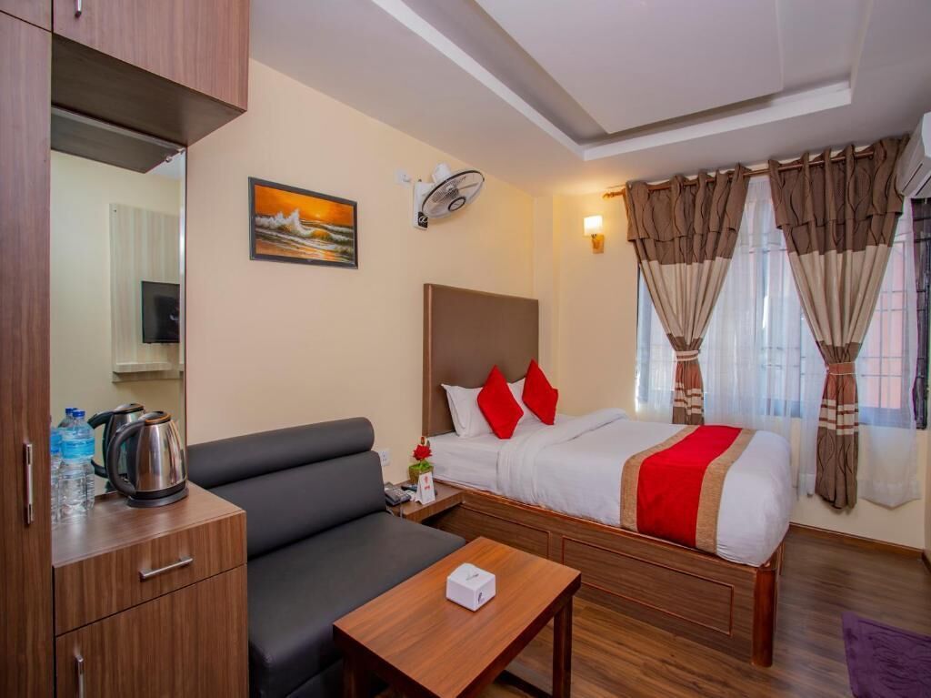 A hotel room with a bed, a sofa, a wooden table, a coffee maker, and a wall-mounted fan.