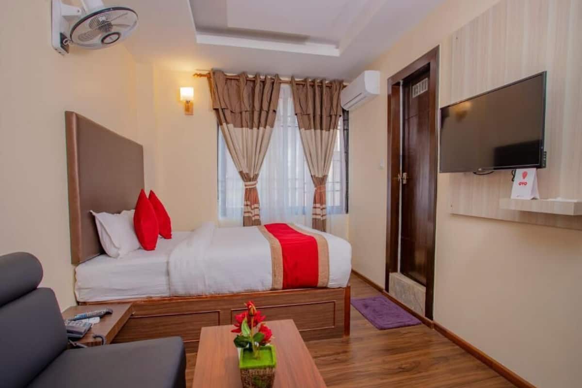 Ein Hotelzimmer mit Bett, Wandfernseher, Nachttisch mit Blumenvase und Deckenventilator.