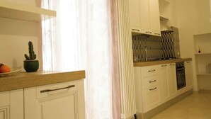 Une cuisine avec des meubles blancs, un plan de travail en bois, un four encastré et une fenêtre ornée de rideaux légers.