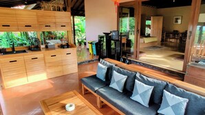A modern, open-plan living area with a wooden sofa, a coffee table, and a kitchen area with a gas stove and sink. - Private nature estate near Mt Merapi, Yogyakarta, stylish villa 4/6 persons (Kabupaten Sleman)