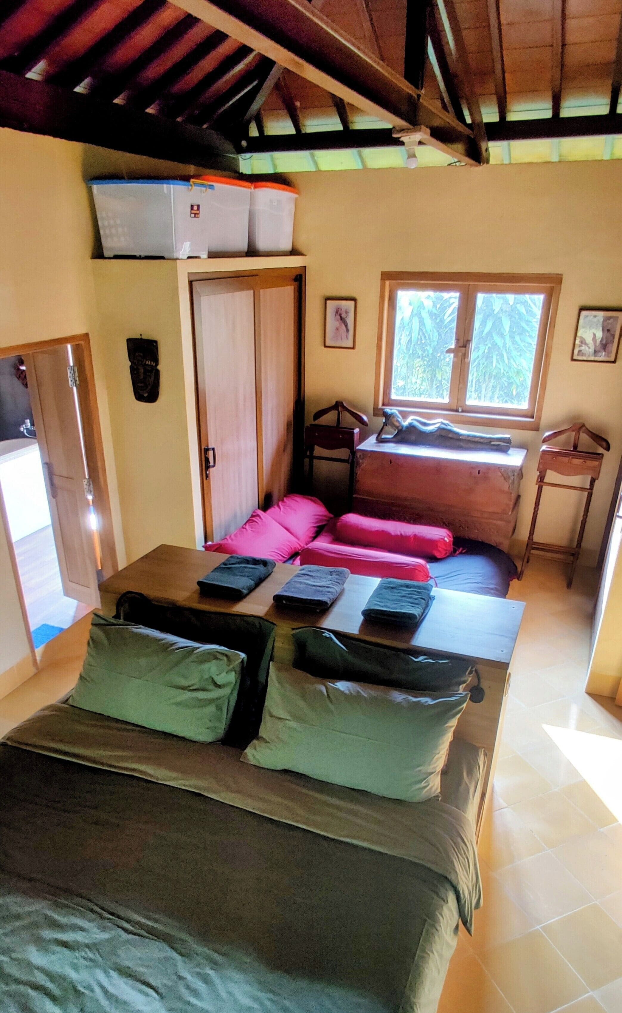 A room with a bed, a window with a view of greenery, a door, and a small table with a chair.