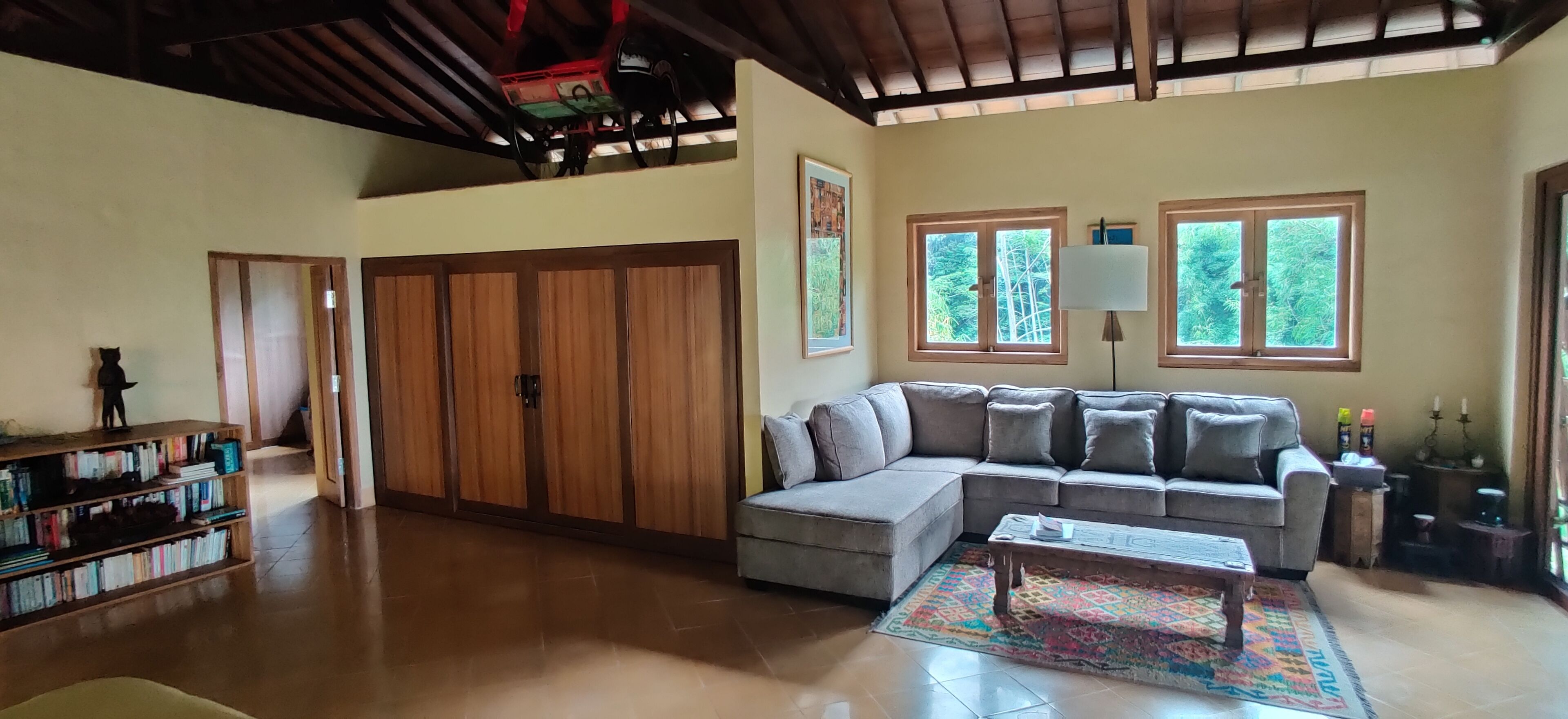 A spacious living room with a large sectional sofa, a coffee table, and a bookshelf. There are two windows providing natural light, and a ceiling fan is visible.