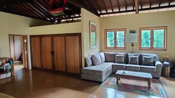 A spacious living room with a large sectional sofa, a coffee table, and a bookshelf. There are two windows providing natural light, and a ceiling fan is visible.