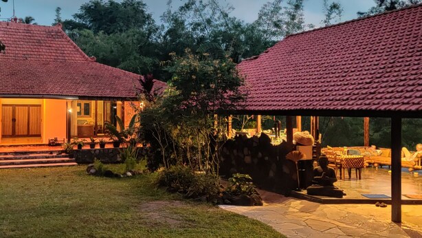 A two-story house with a tiled roof, a stone pathway leading to the entrance, and a covered outdoor seating area with cushions and a statue. - Private nature estate near Mt Merapi, Yogyakarta, stylish villa 4/6 persons (Kabupaten Sleman)