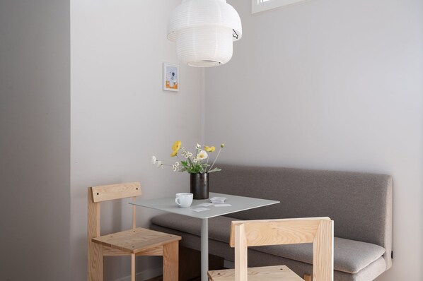 A minimalist room with a grey sofa, two wooden chairs, a small table with a white mug and a vase of flowers, and a white pendant light. - South Studios 2BR by Daniel & Jacob's (København)