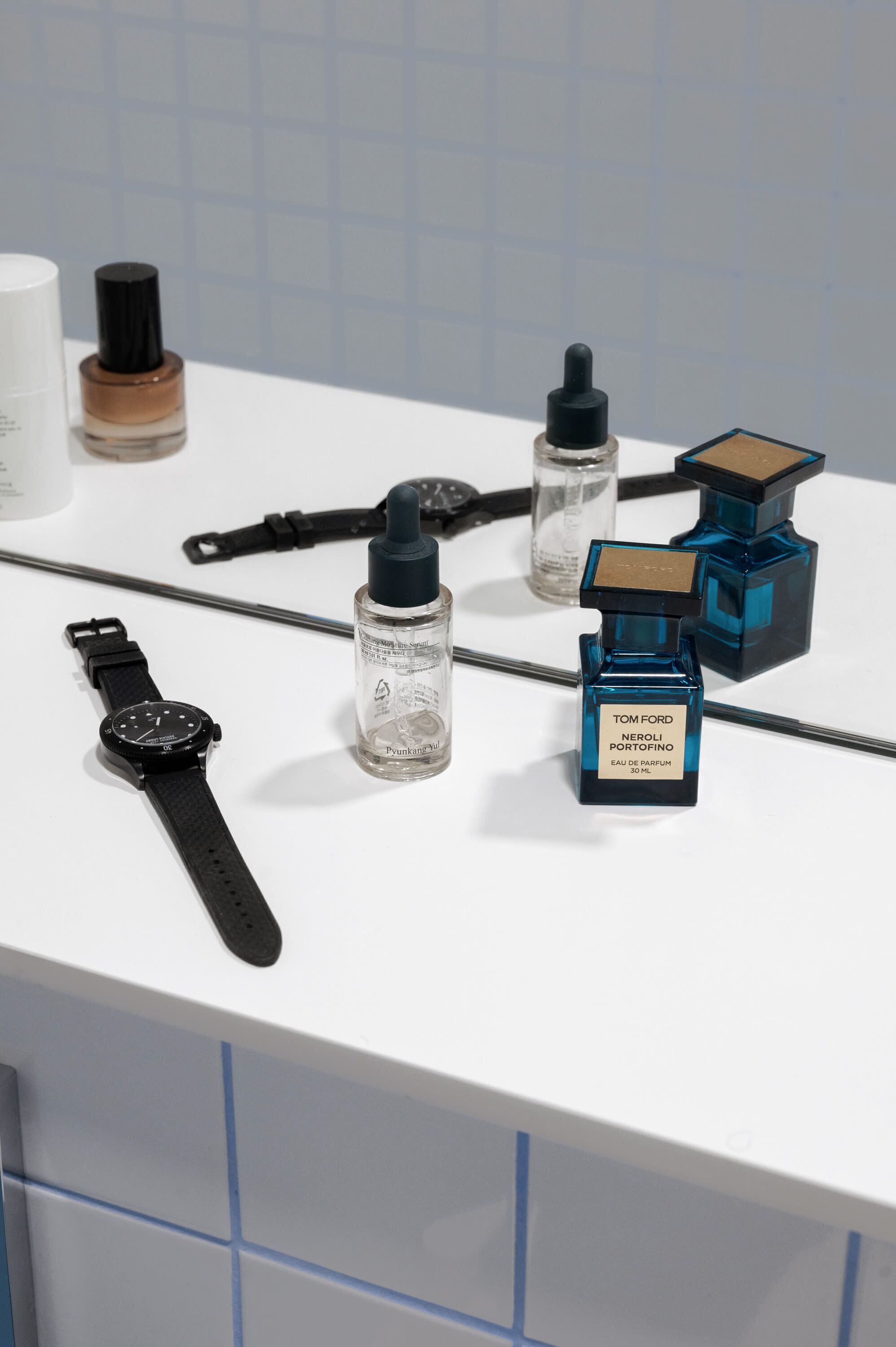 A bathroom counter with various toiletries including a watch, bottles of liquid, and a perfume bottle.