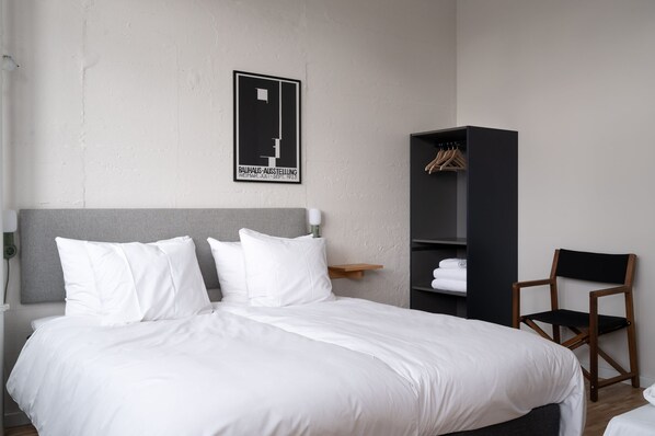 A neatly made bed with white linens, a wooden chair, a black wardrobe, and a wall-mounted coat rack.