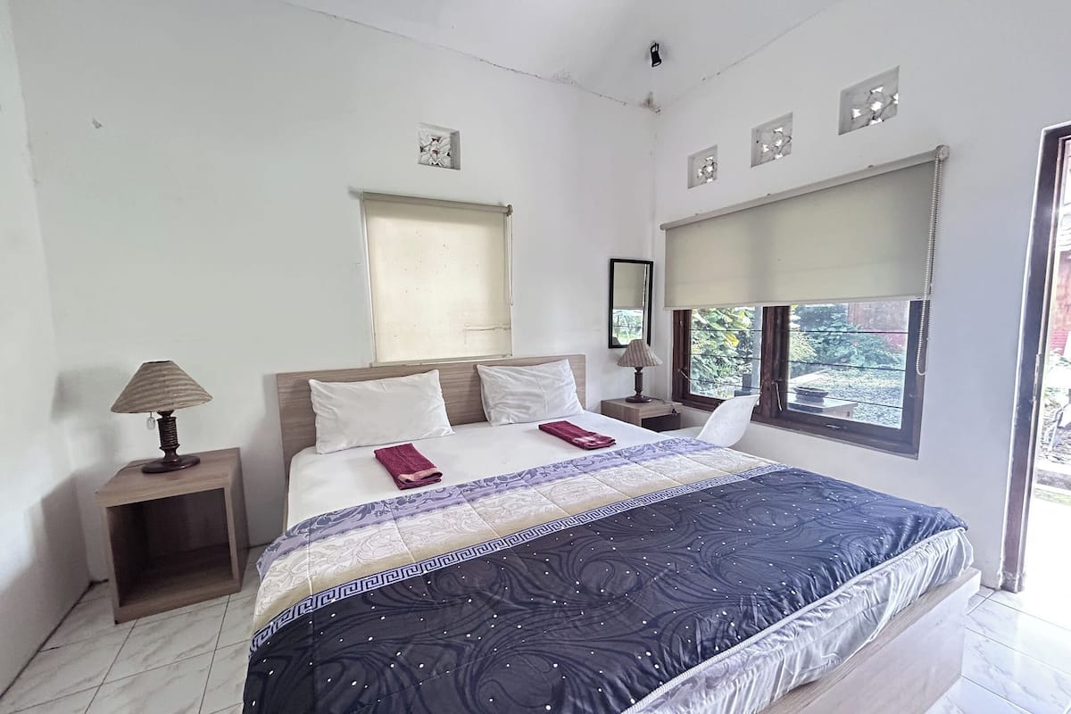A bedroom with a bed, a nightstand with a lamp, a window with blinds, and a view of greenery outside.