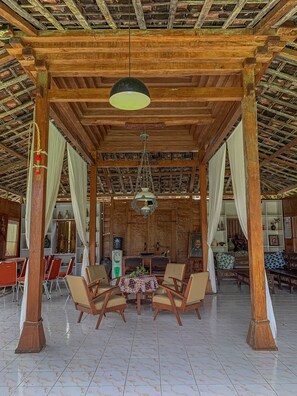 A covered outdoor seating area with wooden pillars, a tiled floor, and a central table with chairs. - Carla Garden (Pakem)