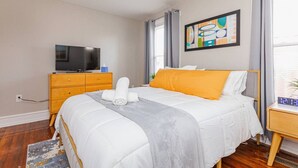 A hotel room with a bed, a TV, a wooden dresser, and a painting on the wall. - Royal Respite: Green Kings & Grey Queens Hideaway! (Richmond)