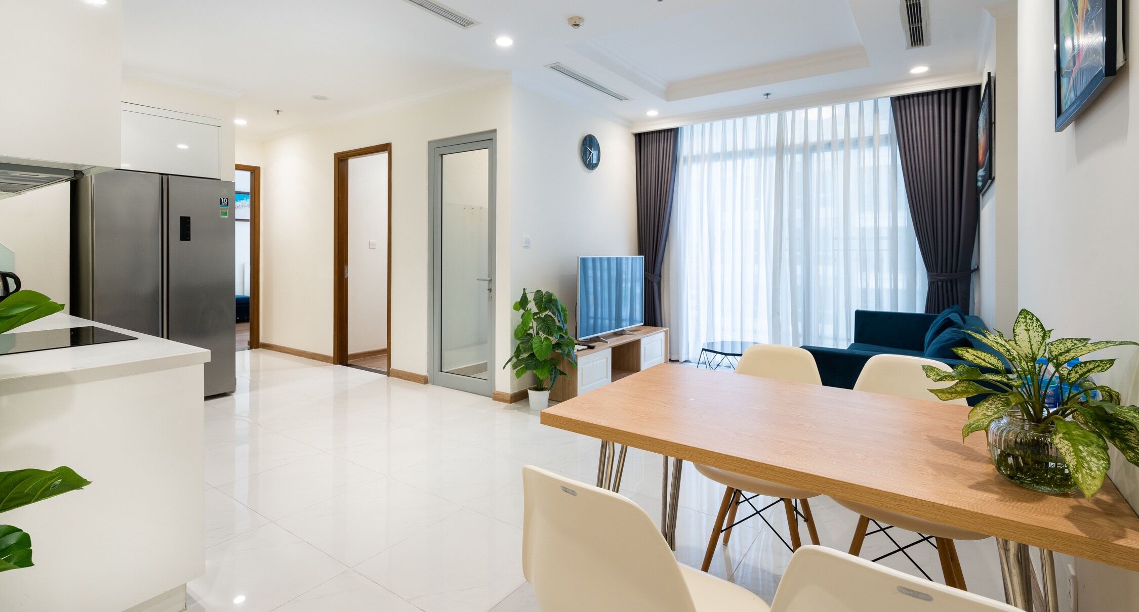Una cocina moderna con refrigerador empotrado, un comedor con mesa y sillas de madera, y una sala de estar con sofá y televisor.