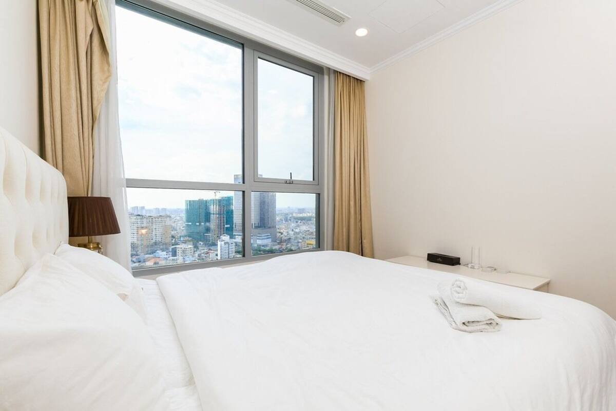 A hotel room with a large bed, a nightstand, a lamp, and a view of a cityscape through a large window.