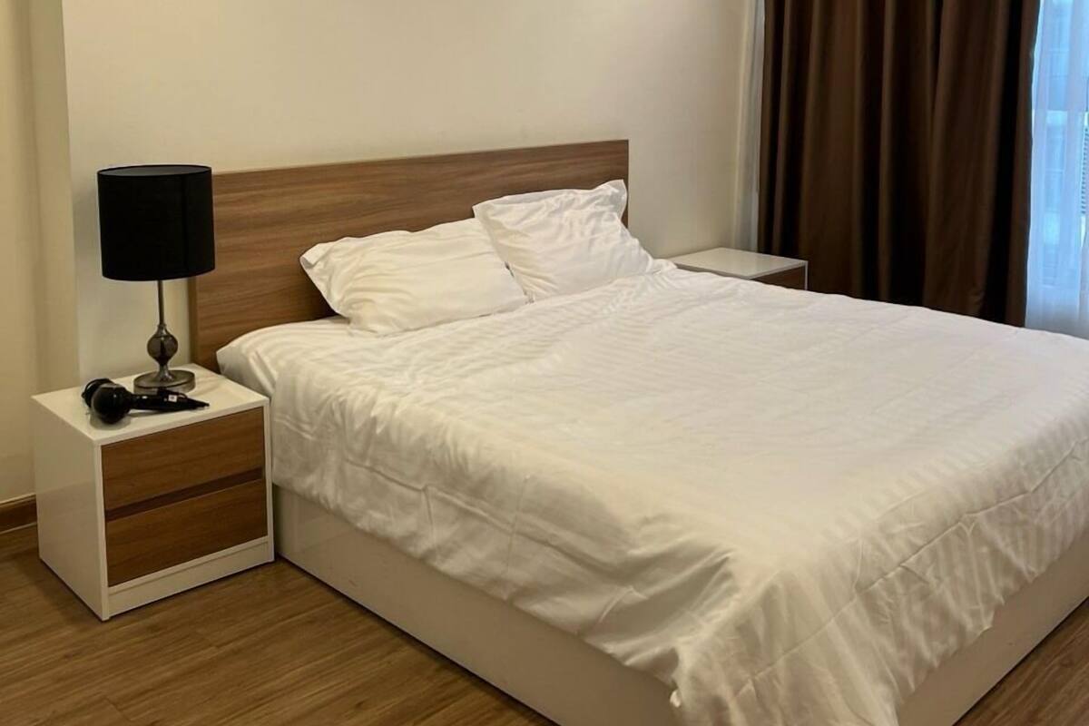 A neatly made bed in a hotel room with a wooden headboard, a bedside lamp, and a nightstand with a lamp and a camera.